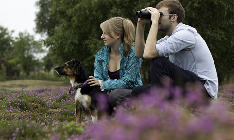 A young man and woman sit amongst flowering heather, with a dog on a lead sat beside them. The woman has her hands on the dog, whilst the man looks through a pair of binoculars.