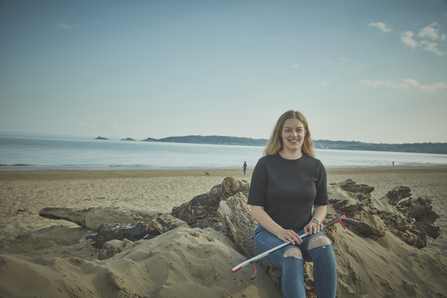 A woman sits on a beach with the sea in the background, holding a litter picker, smiling at the camera