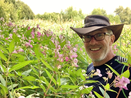Green foliage with small pink flowers fill the image with grassland in background. Man in a brown hat and blue t-shirt next to plants, at chest height