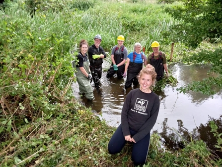 A group of volunteers wearing hard hats, gloves and waders standing in a section of river and a lady wearing a Herts and Middlesex Wildlife Trust T-shirt