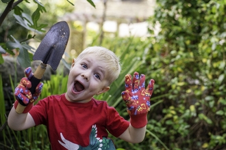 Young boy in red t-shirt with blonde hair wearing gardening gloves holding a spade