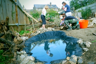 Small pond in foreground, fencing on left and people in background working at ground level to create a garden