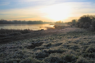 Photograph taken as the sun is rising looking out towards a lake over frosty grass on a winter's morning.