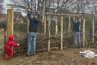 Two adults are lifting fallen tree branches into a wooden frame to form a hedge