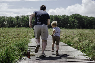 A father and his son running away from the camera down a wooden track, with fields on either side, towards a line of trees