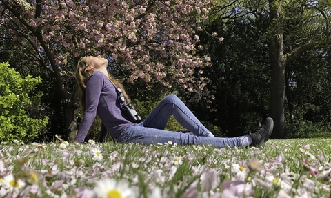 A beautiful young woman sits on the grass in a park, with daisies in the foreground and blossoming trees behind. She has her head thrown back and her eyes closed, enjoying the peace of nature