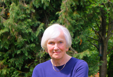 Woman from shoulders up with short grey hair in purple jumper in front of green trees