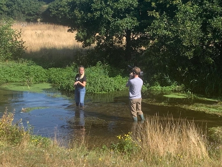Woman standing in stream with cameraman surrounded by trees and grassland