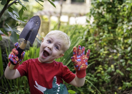 Young boy in red t-shirt with blonde hair wearing gardening gloves holding a spade