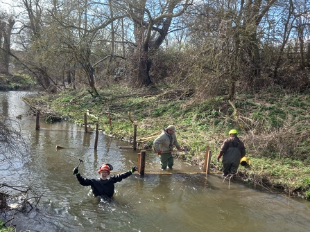 3 volunteers wearing waders and hard hats are standing in a section of river constructing wooden barriers