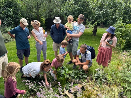 Adults and children outside on a summer walk looking at a pond surrounded by green grass and leafy trees