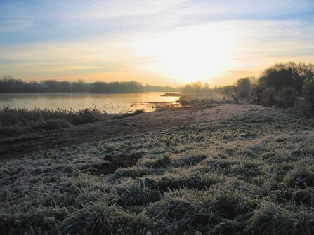 Photograph taken as the sun is rising looking out towards a lake over frosty grass on a winter's morning.