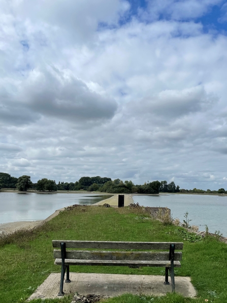 A wooden bench on a grassy prong looking out over the divide between bodies of water on a cloudy day. In the distance, the opposite banks are lined with trees.