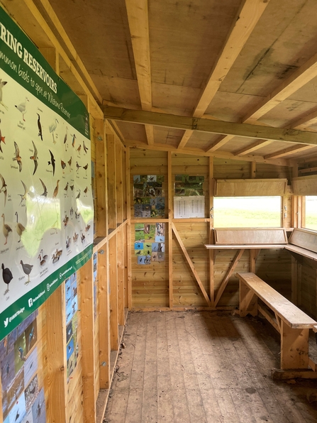 The interior of a wooden shed-like building with a long bench and windows cut into the side to look out over the reservoir. On the back wall are posters and photos detailing a variety of bird species that may be spotted.