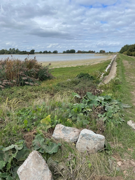 A view of a well-trodden grass pathway around a large, irregularly shaped reservoir. The blue water is quite low and does not fully reach up the banks.