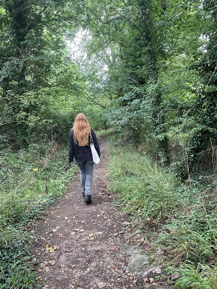 A dirt path leading through a woodland of straightish medium sized trees. Anna is walking down the path she is wearing a black fleece, jeans and walking boots.