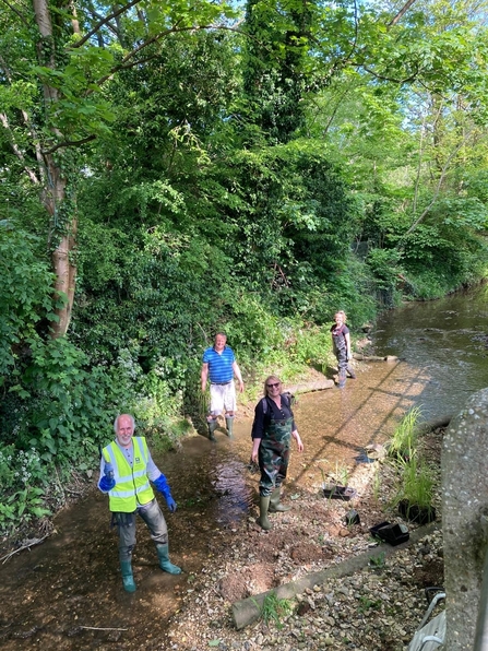2 men wearing wellies and 2 women wearing aders are standing in a shallow river. One bank is lush and green with trees and vegetation and the although the other half is currently more bare, there are potted native plants laid out on top of the soil ready to be planted.