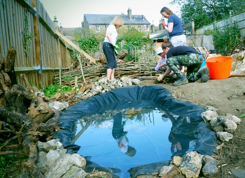 Small pond in foreground, fencing on left and people in background working at ground level to create a garden