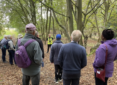 A group of people wearing outdoor clothes gathered in a clearing in a forest in early autumn listening to a man in a reflective jacket who is giving a talk