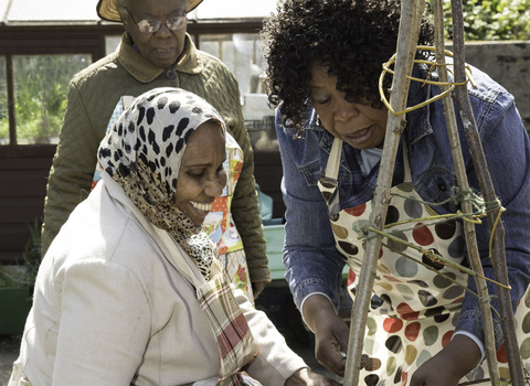 3 ladies gathered around a raised bed tying strips of thin branch to a tripod of thicker branches to create a frame for plants to grow up.