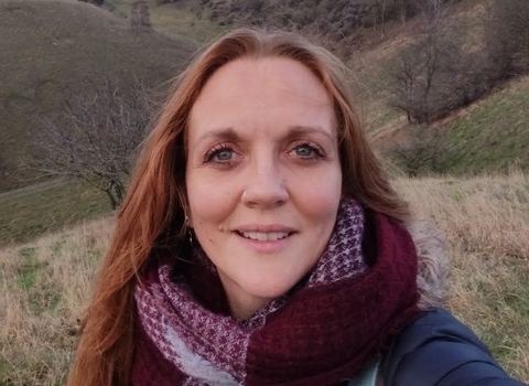 Amy looks at the camera with a hilly late winter landscape behind her